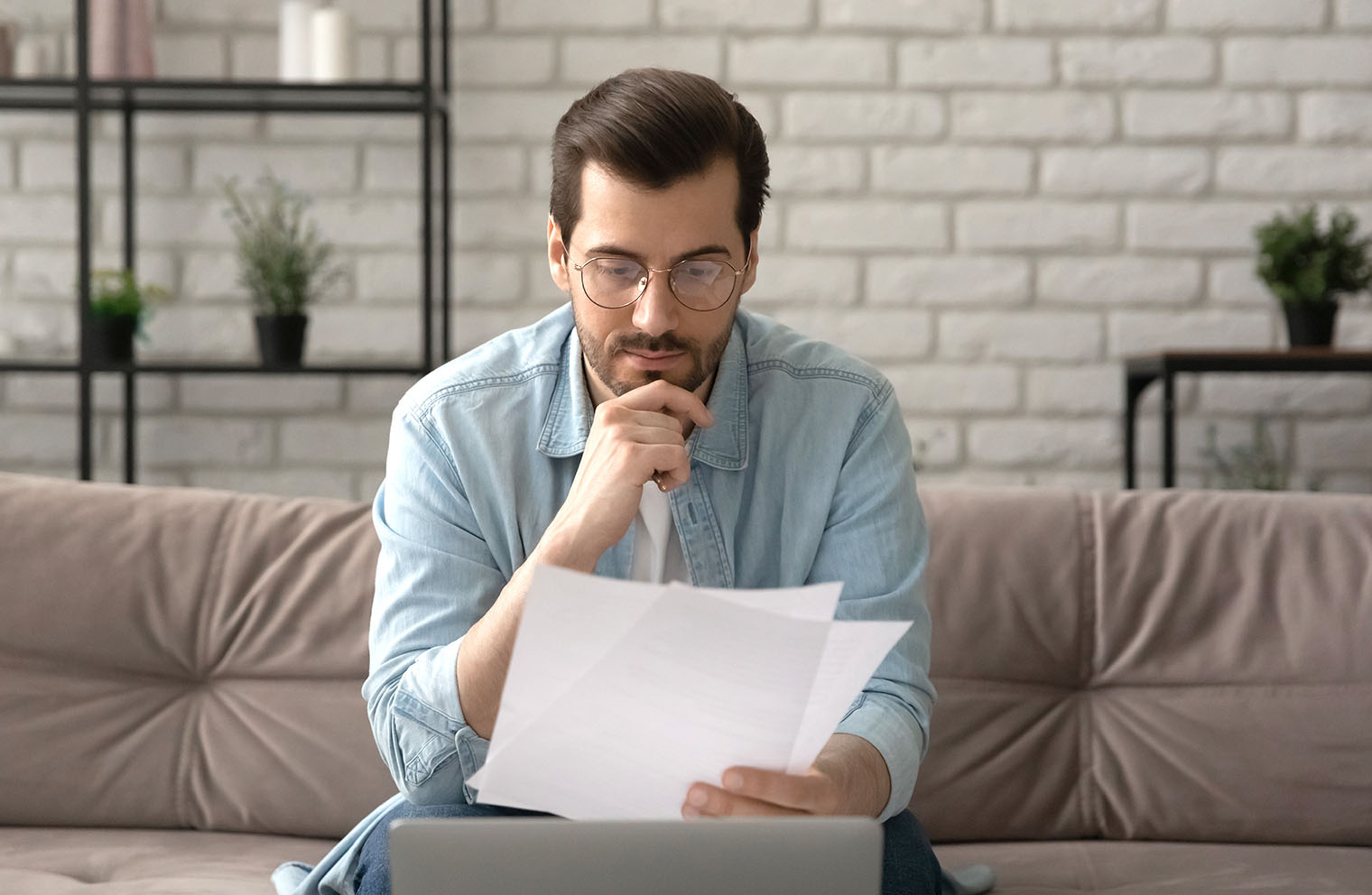 Image of a man sitting on a couch reading sheets of paper with a laptop in front of him