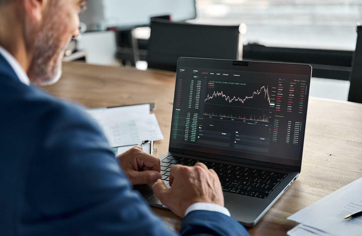 A man in a suit sitting in front of a laptop reading stock market information