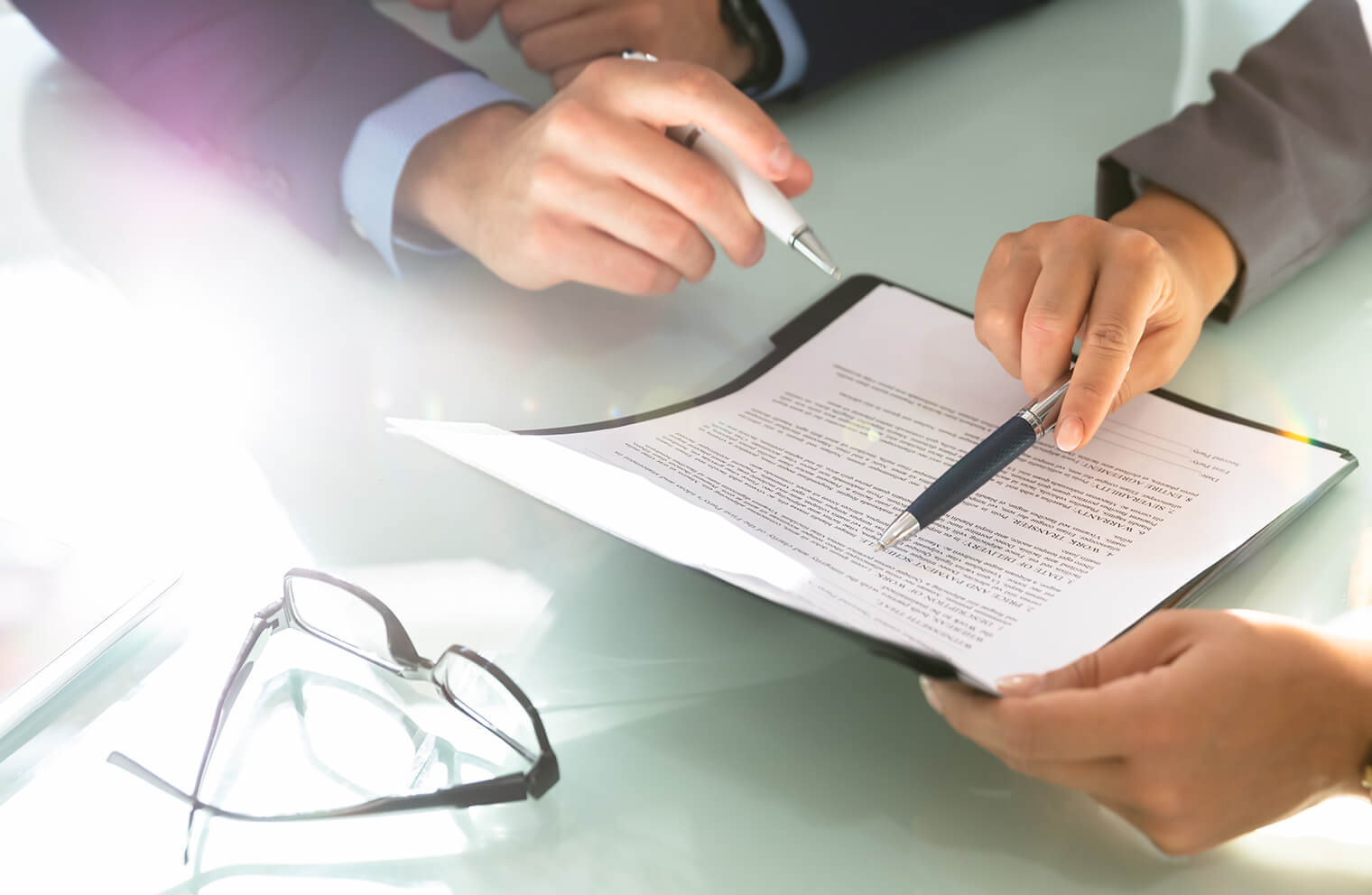 Close up picture of two people reviewing a document using their pens on a desk with glasses on the desk.