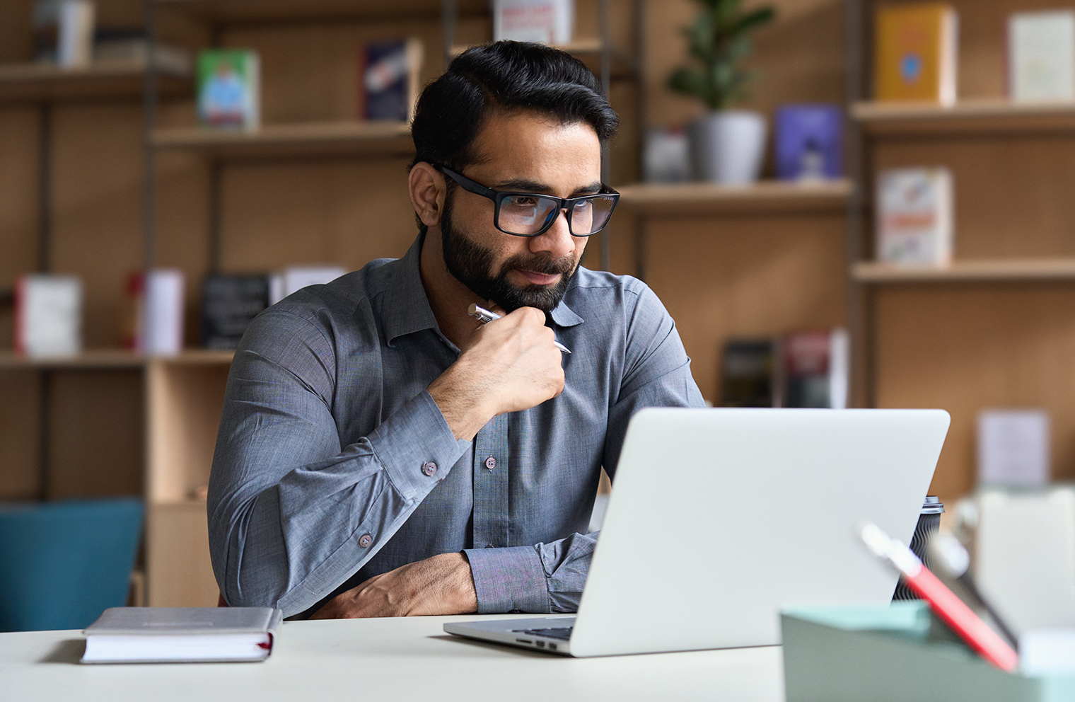 A man thoughtfully sitting in front of a laptop with a pen and notebook.