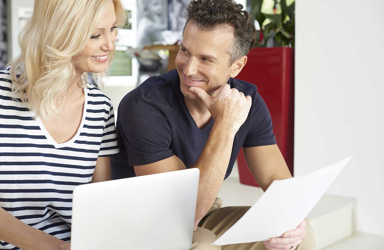 Picture of a couple smiling at each other while sitting on a couch and holding papers.