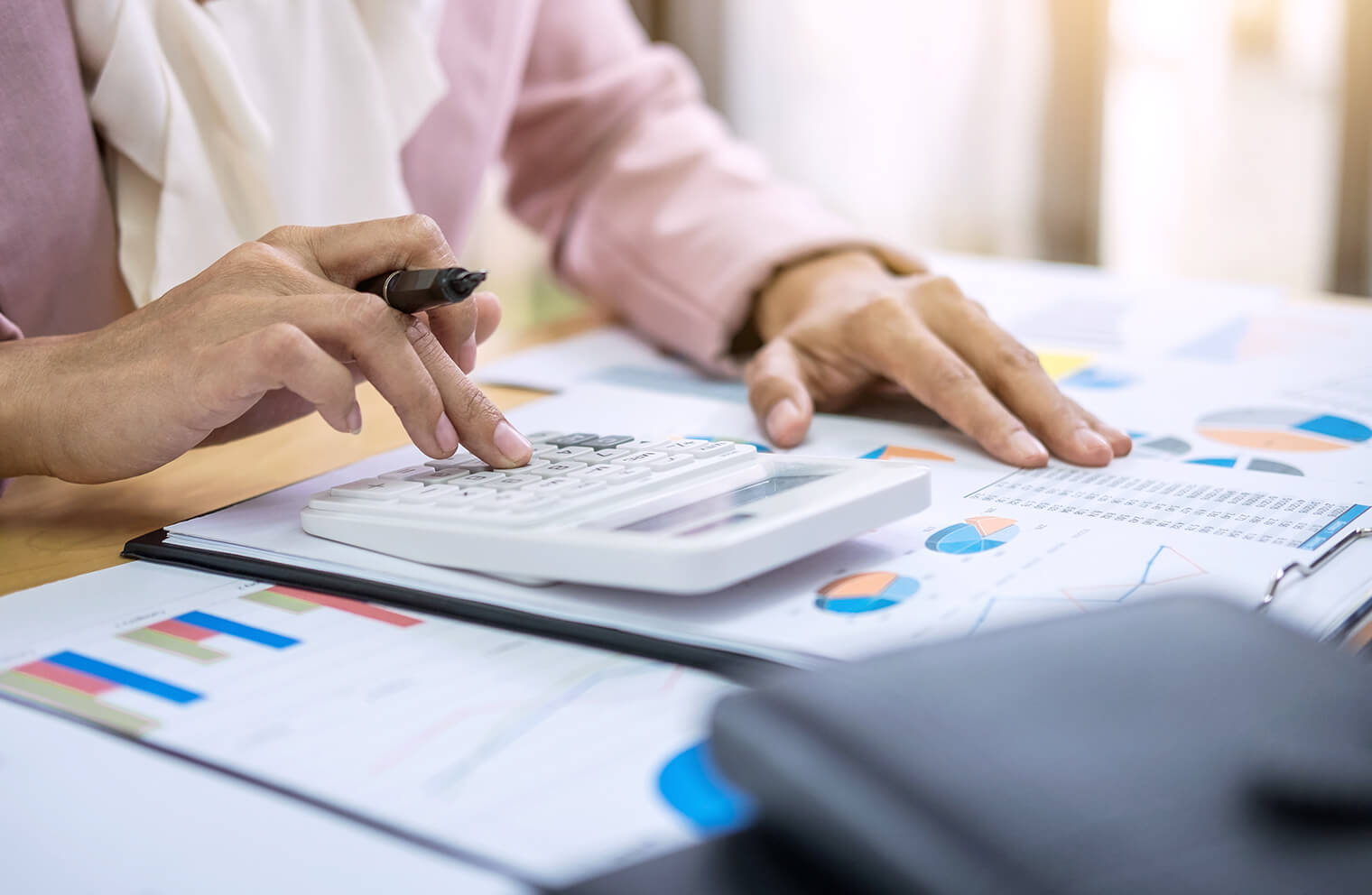 Close up picture of a woman in a pink blazer  using a calculator while reviewing and looking at financial statements with pie charts and graphs.