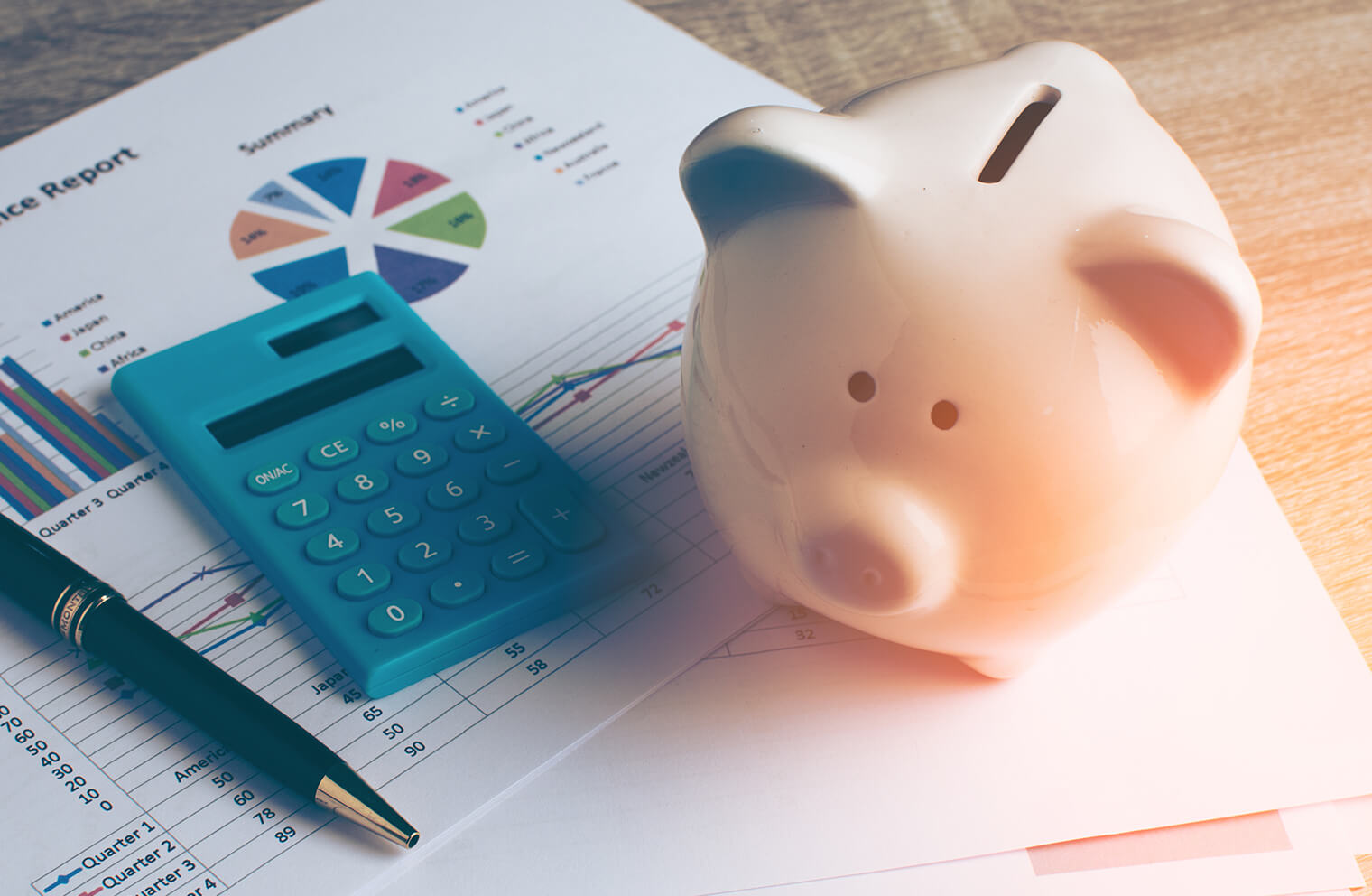 Close up image of a piggy bank, blue calculator, and pen on top of a finance report with graphs and charts.