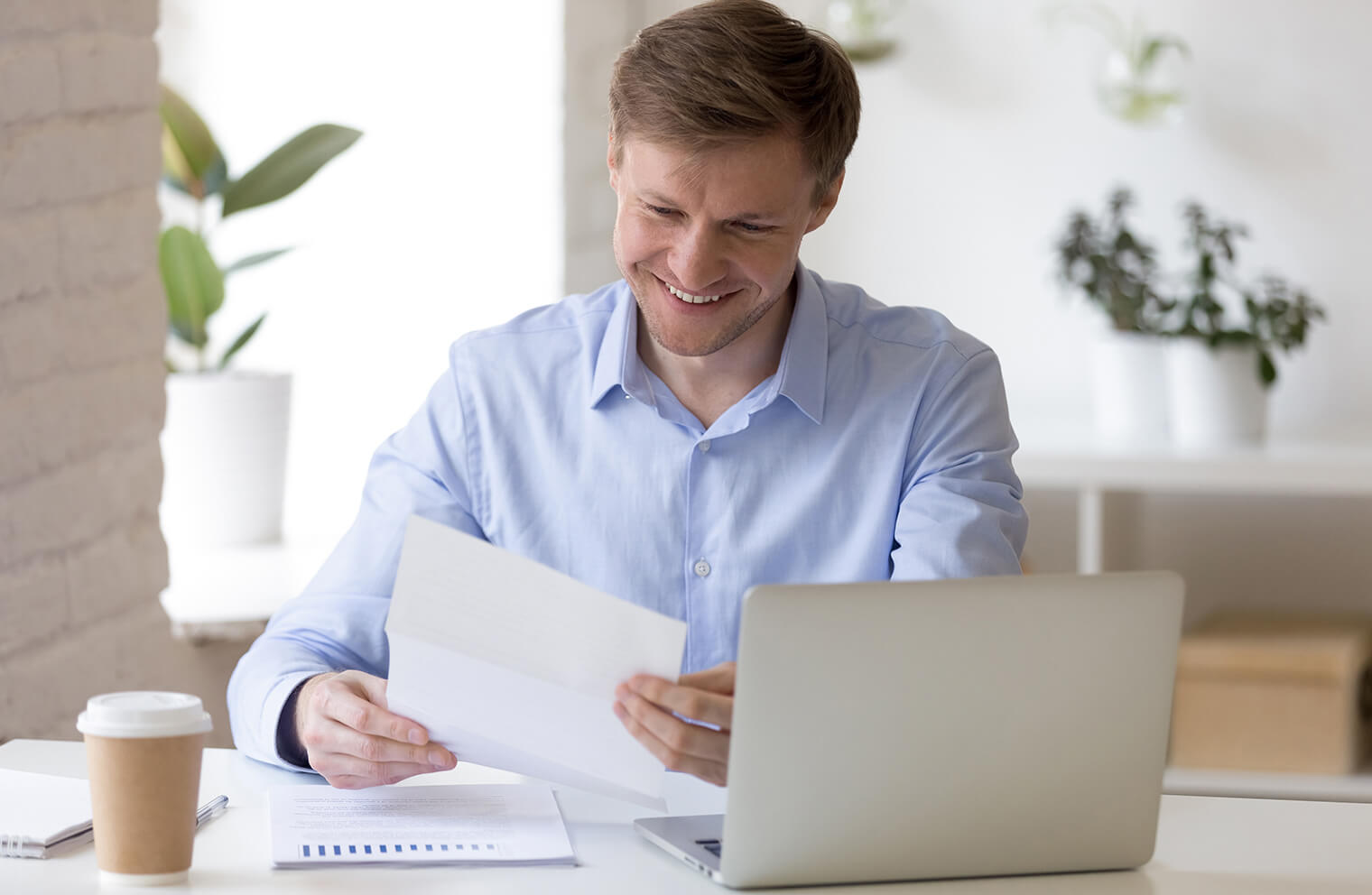 Picture of a man in a blue dress shirt, smiling and holding a piece of paper. He is sitting at  a white desk that has notebooks, coffee, documents, and a laptop on it.