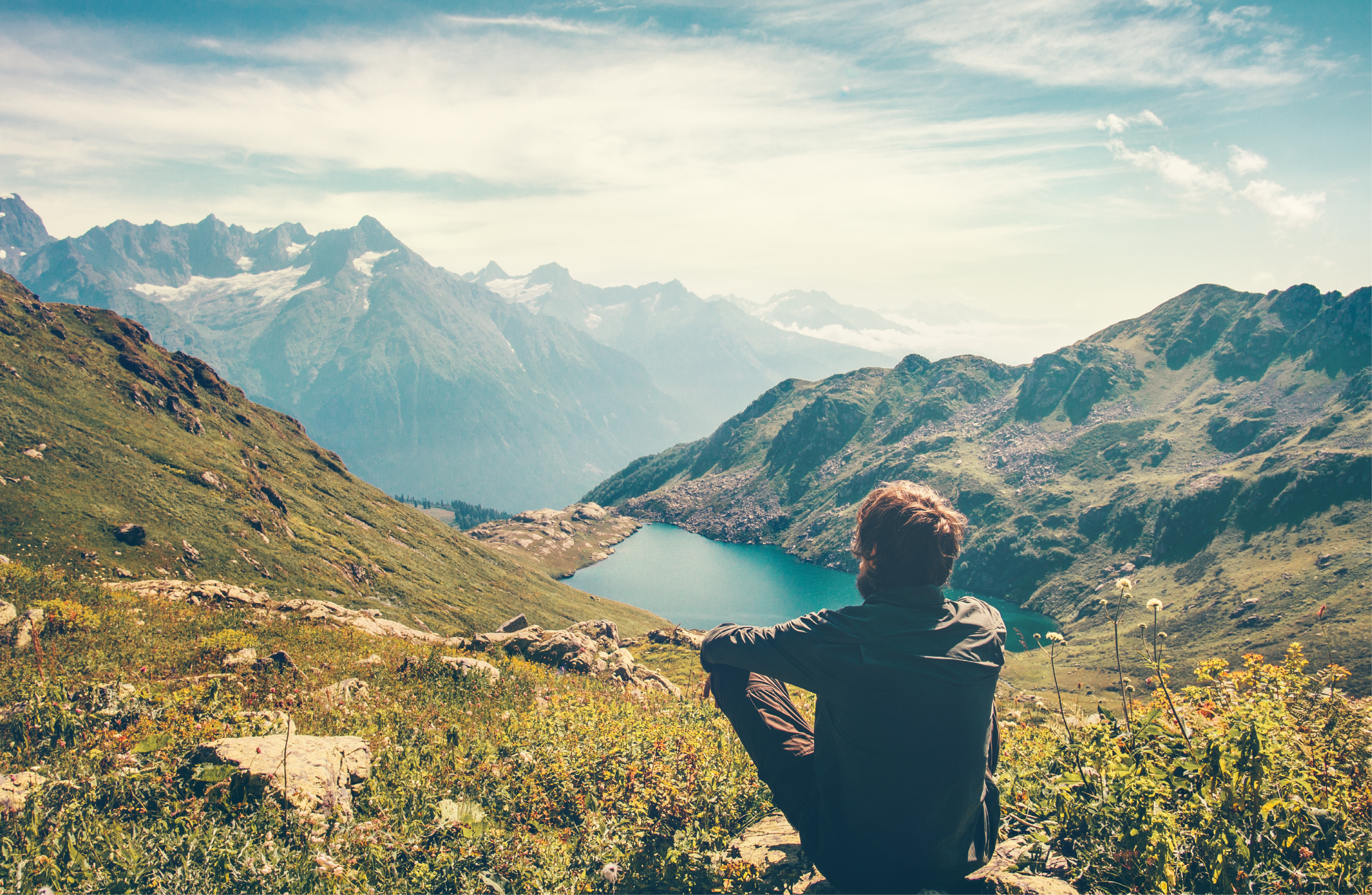 A pastor overlooks majestic mountains and a lake as he sits on a mountaintop. 